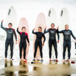 Deelnemers aan de Olympische Spelen voor strand en water opgesteld op het strand in surfuitrusting en met een surfplank